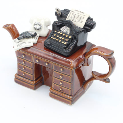A whimsical ceramic desk figurine with a handle and drawer details, decorated with a black typewriter, a white rotary phone, and papers on top, photographed on a white background.