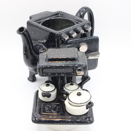 A black speckled Paul Cardew ceramic novelty teapot  designed to look like a stove, with three small white pots on a black burner base, photographed on a white background.