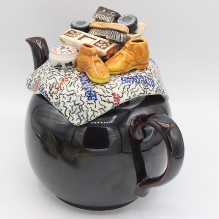 A ceramic teapot with a glossy black body and a patterned lid featuring miniature figurines, including two orange boots, a small white pot, and a stack of books and hats, photographed on a light gray background.