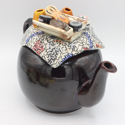 A glossy dark brown ceramic teapot with a patterned fabric ceramic lid and a whimsical miniature cargo load of shoe shine pieces resting on top, photographed on a light gray background.