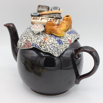 A glossy black ceramic teapot with a handle and spout, decorated with a whimsical lid design featuring a patterned cloth, miniature show shine brushes, and a pair of orange boots, photographed on a light gray background.