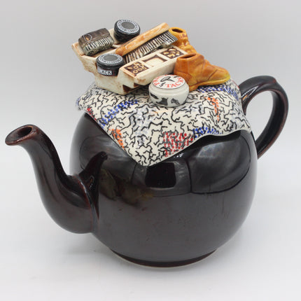A novelty ceramic teapot with a glossy dark brown body, decorated with a patterned fabric lid insert and topped with miniature shoe iems.
