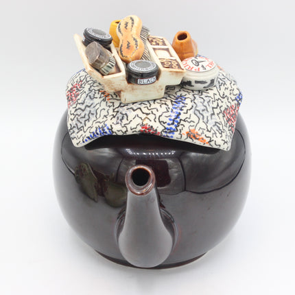 A glossy dark brown ceramic teapot with a highly detailed, hand-painted miniature tea party scene on top of its lid. The scene features small ceramic cups, saucers, a teapot, and a patterned napkin on a clean white background.