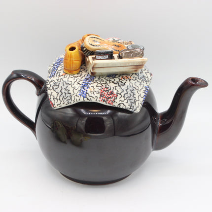 A dark brown glossy ceramic teapot with a lid, decorated with a colorful floral and geometric patterned fabric top and small decorative figurines including a yellow teapot and a miniature book.