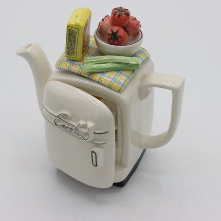 A ceramic teapot shaped like a vintage cream-colored refrigerator, with the door open to reveal a small compartment. On top of the teapot sits a yellow-and-white plaid cloth holding a yellow butter-like block, green beans, and a small bowl of red tomatoes.