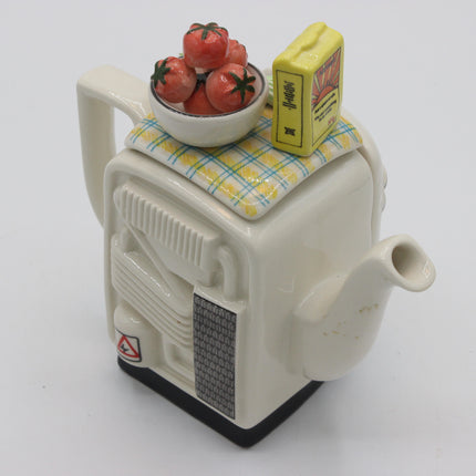 A novelty ceramic teapot designed to look like a vintage appliance, with a small bowl of red tomatoes and a yellow matchbox on top, photographed on a white background.