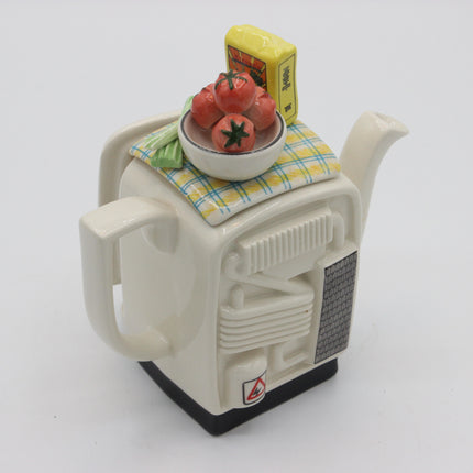 A novelty ceramic teapot designed to look like a retro computer or appliance, in off-white with a black base. The top is decorated with a yellow-and-blue checkered cloth, a small bowl of red tomatoes, and a yellow packet.