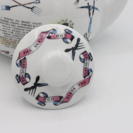 A close-up of a white ceramic lid featuring pink and blue decorative ribbons with the words 'GOD BLESS THE GARDENER' and black hand-drawn garden motifs, with part of the matching planter body visible behind it.