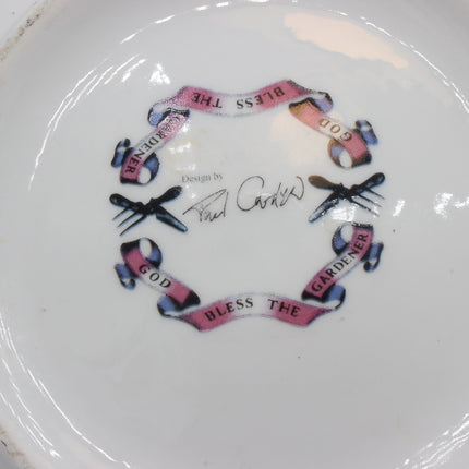 A close-up of the bottom of a white ceramic plate featuring a printed circular design with pink banners reading 'God Bless the Gardener' , and a signature that reads 'Design by Paul Cardew'.