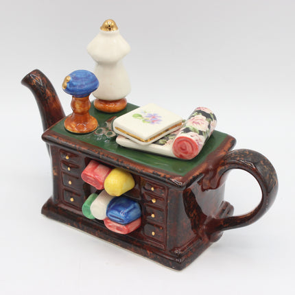 A vintage ceramic sewing cabinet figurine with a handle, featuring a green top, brown drawer base, and miniature sewing accessories including a white dress form, floral fabric rolls, and colored spools.