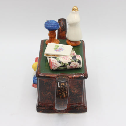 A vintage ceramic jewelry box shaped like a desk or cabinet, featuring a dark brown speckled body, a green top, and miniature decorative figurines on top including a white dress, blue and brown mushroom shapes, and a floral-covered rolled scroll.