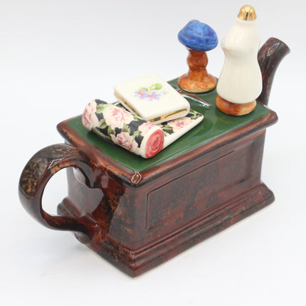 A ceramic figurine of a sewing machine with a dark brown speckled body, a green top, and decorative items including a floral ribbon, a small floral card, and two small figurines on top, set against a plain white background.