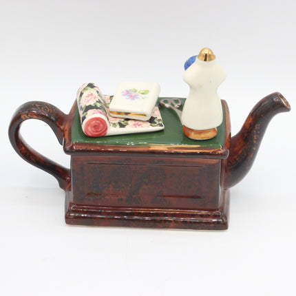 A decorative teapot-shaped box with a glossy dark brown body, a green top surface, and miniature sewing-themed accessories including a floral fabric roll, a small floral notebook, scissors, and a cream dress form on a white background.