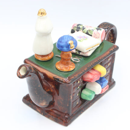 A ceramic teapot with a green top, brown body, and colorful miniature accessories including a white dress form, a blue-and-gold mushroom-shaped ornament, a floral fabric roll, and small colored stones in the side compartment, photographed on a white background.