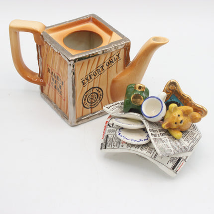 A ceramic novelty teapot shaped like a wooden crate with 'EXPORT ONLY' text, shown on a white background with small decorative accessories including a miniature green toy camera, white cup, yellow bear figurine, and newspaper folds.