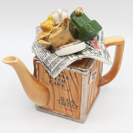 A novelty ceramic teapot shaped like a cardboard shipping box with a spout and handle, topped with a stack of newspaper and small figurines including a green suitcase and a yellow duck.