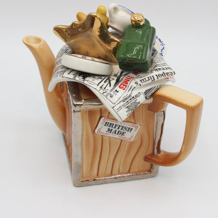 A ceramic novelty teapot shaped like a wooden crate with a handle, spout, and lid, decorated with miniature luggage items and newspaper, featuring a 'BRITISH MADE' label on the front, set against a plain white background.