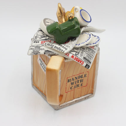 A ceramic novelty mug shaped like a wooden crate with a handle, decorated with newspaper cutouts and small figurines including a green frog and a gold bird, on a white background.