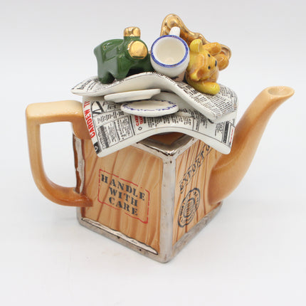 A novelty ceramic teapot shaped like a wooden crate with 'HANDLE WITH CARE' printed on the front. The teapot features a glossy amber-brown finish, an open newspaper lid, and whimsical figurines including a green pig, a yellow teddy bear, and a small white cup with blue trim resting on top.