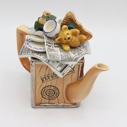 A ceramic teapot shaped like a wooden crate with a handle, spout, and lid, decorated with newspaper print and topped with small figurines including a yellow teddy bear, a white cup, and a green hat, photographed on a white background.