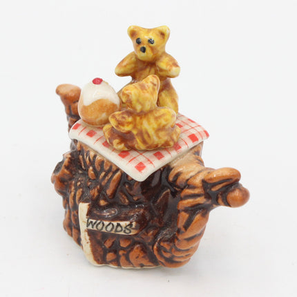 A small ceramic figurine of three teddy bears gathered around a red-and-white checkered blanket on top of a brown textured base, photographed on a plain white background.