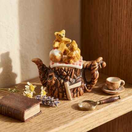 A miniature ceramic teapot scene figurine featuring two small teddy bears sitting on a red-and-white checkered blanket atop a brown tiger-shaped base, with a small white cake on top, photographed on a plain white background.