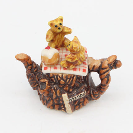 A miniature ceramic tea-time scene figurine featuring two small teddy bears sitting on a red-and-white checkered blanket atop a brown tiger-shaped base, with a small white cake on top, photographed on a plain white background.