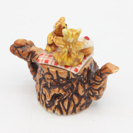 A small decorative figurine shaped like a tree trunk or pot with two small teddy bears sitting on top of a red-and-white checkered blanket, photographed on a plain white background.