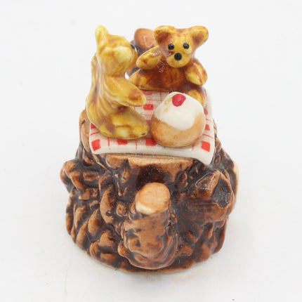A small ceramic figurine of two teddy bears sitting on a log, sharing a cupcake with a red and white checkered blanket, set against a plain white background.