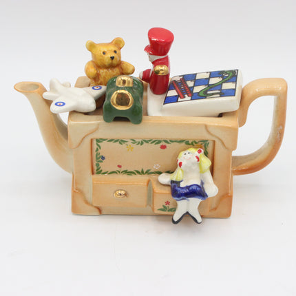 A whimsical ceramic teapot figurine styled like a tea cabinet, featuring a teddy bear, a toy soldier, a rabbit, a green toy car, a chess board, and a small Alice-in-Wonderland figure on a white background.