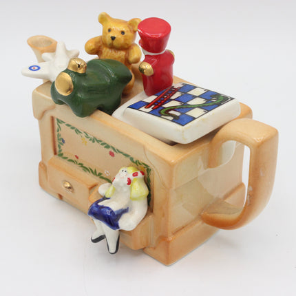 A vintage ceramic novelty tea pot shaped like a toy box, decorated with small figurines including a teddy bear, a red soldier, a green frog, a chess piece, and a seated girl figure, shown on a white background.