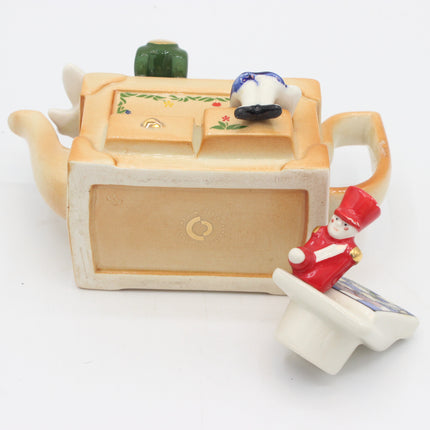 A vintage ceramic novelty teapot with a drawer front and a small figurine of a child in a red uniform sitting on the base, set against a plain white background.