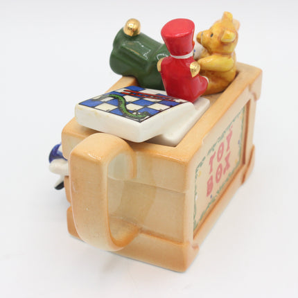 A vintage ceramic toy box teapot with a handle, decorated with a green frog, a red figure, a yellow teddy bear, and a checkered box on top, with 'TOY BOX' written on the side.
