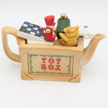 A ceramic toy box shaped like a teapot, with the words 'TOY BOX' on the front. The top of the box is filled with small toy-like characters and objects, including a red-and-white striped figure, a green figure, a yellow bear figure, and a white rabbit figure, all photographed on a plain white background.