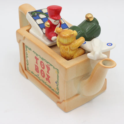 A vintage ceramic toy box teapot featuring toy characters and toys, including a red soldier figure, a green toy train, a yellow teddy bear, and a white airplane, on a white background.