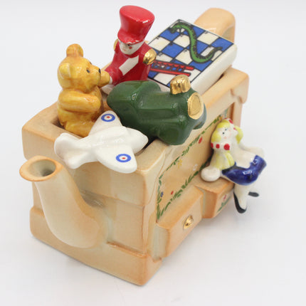 A ceramic novelty teapot shaped like a toybox, filled with small toy-like figurines including a yellow teddy bear, a red-and-white soldier, a green frog, a white airplane, and a white-and-blue cat, photographed on a white background.