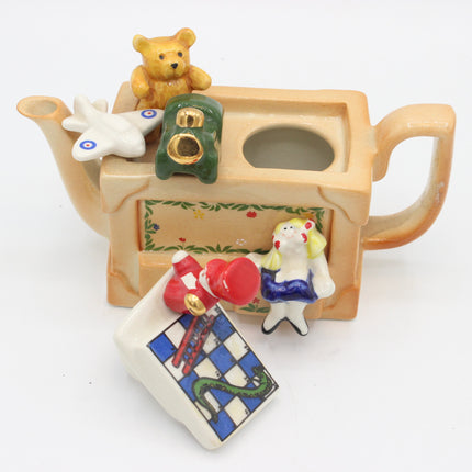 A decorative ceramic teapot-shaped container with a handle and spout, decorated with small figurines including a teddy bear, a white bird, a green hat, a white rabbit, and a chess-piece-themed lid placed in front on a white background.