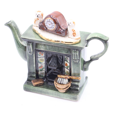 A ceramic teapot shaped like a fireplace, in a muted green glaze, featuring a miniature clock, two small dogs, floral window panels, and a small gold-toned bowl on a white background.