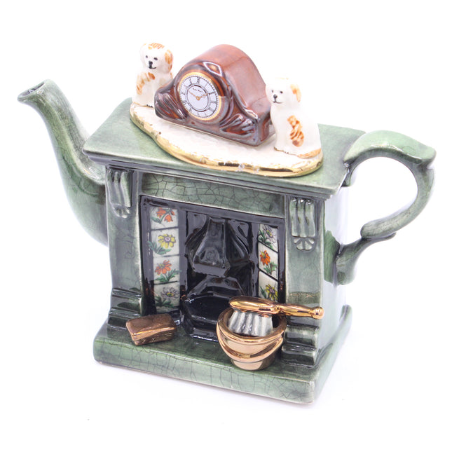 A ceramic teapot shaped like a fireplace, in a muted green glaze, featuring a miniature clock, two small dogs, floral window panels, and a small gold-toned bowl on a white background.