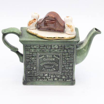 A ceramic teapot shaped like a green brick house with a small brown house and two white birds on top, photographed on a plain white background.