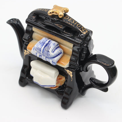 A novelty ceramic tea towel holder shaped like a miniature black teapot, featuring a gold dog topper, gold lettering, and a miniature blue-and-white patterned towel.