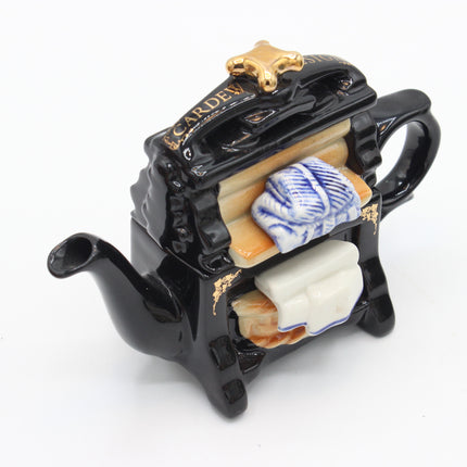 A novelty ceramic tea towel holder shaped like a black teapot, featuring gold lettering, a gold star handle, and a cutaway design showing blue-and-white and cream tea towels inside.