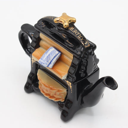 A miniature black ceramic teapot shaped like a coffin, featuring gold accents, a blue and white striped pillow, and a beige quilted blanket, displayed on a clean white background.