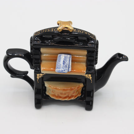 A glossy black ceramic teapot shaped like a vintage typewriter, featuring gold lettering on the lid, a gold handle, a gold spout, and a beige and blue paper roll detail on the body, set against a plain white background.