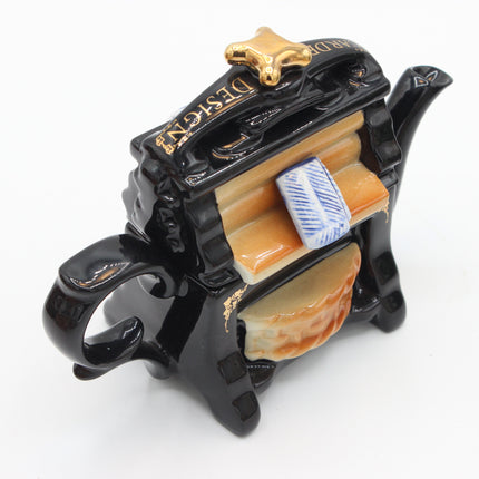 A decorative ceramic teapot shaped like a coffin, finished in glossy black with gold lettering and trim, orange-brown interior accents, and a small blue-and-white patterned accent piece, photographed on a white background.