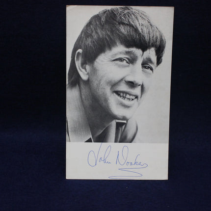 Black and white photo of a smiling John Noakes with a signature below on a dark background