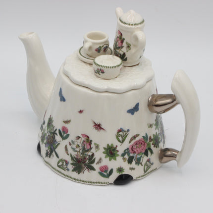 A vintage white porcelain teapot with a floral and butterfly pattern, featuring a matching small teapot and cup set on top, photographed against a plain white background.