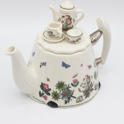 A vintage-style ceramic teapot with a floral and butterfly pattern, shown on a plain white background. The teapot features a white body with pink, green, and blue botanical illustrations, a white lid, a white handle, and a silver metal spout.
