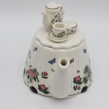 A white porcelain teapot shaped like a flower, decorated with pink roses, green leaves, blue butterflies, and small black and white spotted accents, shown on a plain light gray background.