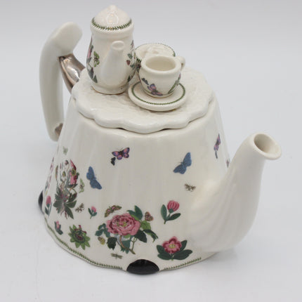 A vintage-style white porcelain teapot with a lid, decorated with pink roses, green leaves, and colorful butterflies, shown with a matching teapot and cup on a saucer.